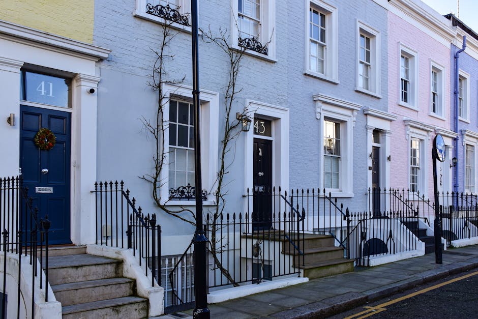 Photograph of a row of three terraced houses on a residential street in Covent Garden during daylight, with the focus on the front entrances. The first house has a blue door decorated with a festive wreath, white painted brick walls, and a small set of concrete stairs leading up to the entrance, bordered by black metal railings. The middle house features a black door with house number 43, white walls, and a small steps leading to the door. The third house has a white door, a pastel pink exterior, and blue accents on the window frames, also accessed via small stairs with black railings. A black sidewalk lamp and a street sign are visible in the foreground, with leafless trees and decorative window railings on the upper floors contributing to the residential street atmosphere. The scene suggests an urban environment suitable for house relocations and furniture transport, with prominent features of the street's architecture and entryways.