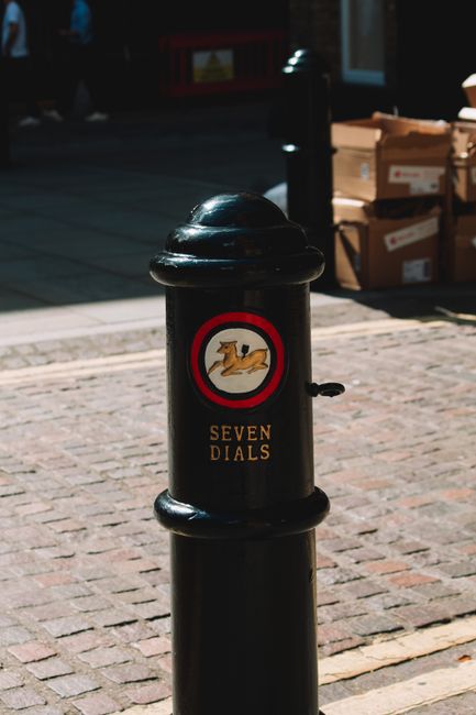 A black street bollard located on a cobbled pavement in a city area, featuring a circular sticker with a graphic of a dog running, and the text 'SEVEN DIALS' below it. Behind the bollard, there are cardboard boxes stacked on the pavement near a building entrance, indicating a packing or loading area typical of house removals and moving logistics. The scene is lit by natural daylight, with a shadow cast on the ground, and the background shows part of a doorway and some outdoor elements, consistent with a busy urban environment that may be subject to parking or loading restrictions affecting furniture transport and home relocation services offered by Man With a Van Covent Garden.