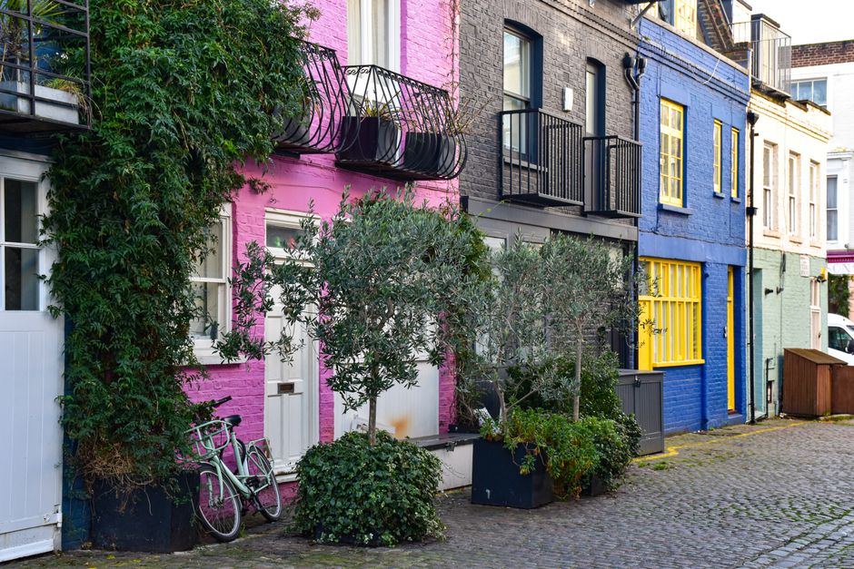 A row of colorful residential buildings in a narrow street, painted in pink, blue, and light green, with small balconies featuring black railings and potted plants. In front of the pink building, there are two green bicycles parked beside a white door. The street surface is cobblestone, and several potted plants and small trees are placed along the pavement, with some greenery climbing parts of the building facades. The scene appears to be part of a home relocation or moving process managed by Man With a Van Covent Garden, illustrating the urban environment where furniture transport and packing activities may take place under Camden Council loading bay regulations.