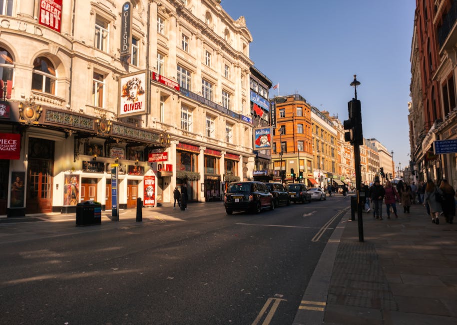 A street scene in Covent Garden during daylight, featuring multi-storey buildings with ornate facades and large windows, some with signs for theaters and shops. The street has a mix of cars, including black and white vehicles, parked along the curb and driving through the intersection. Pedestrians are walking on the sidewalk, and a traffic light is visible at the corner. The environment appears busy yet organized, with clear weather and natural lighting. In the context of house removals and furniture transport, this image illustrates the urban environment where moving services by Man With a Van Covent Garden typically operate, often involving street-level loading and unloading for home relocation projects.