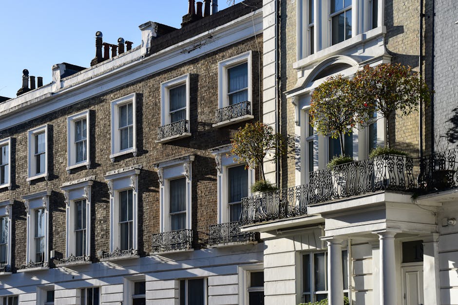 Exterior view of a multi-storey Georgian-style residential building with brick facades, white window frames, and decorative molding. Several windows have small black wrought iron balconies, some with potted plants and small trees. The building's ground floor features large white panels and a white entrance door. Sunlight casts shadows across the facade, highlighting the architectural details. This setting reflects an urban residential area in Covent Garden, where house removals and furniture transport may involve navigating narrow balconies and street access for efficient home relocation services. Occasionally, the process includes packing, loading, and carefully transporting household furniture and belongings from such buildings, with Man With a Van Covent Garden providing professional removals in these settings.