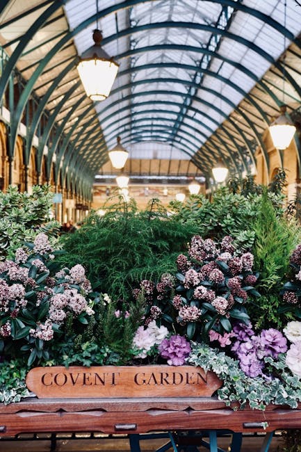 Inside a glass-roofed structure resembling a greenhouse or plant shop, various potted plants and floral arrangements cover a wooden cart labeled 'COVENT GARDEN.' The plants include leafy green foliage