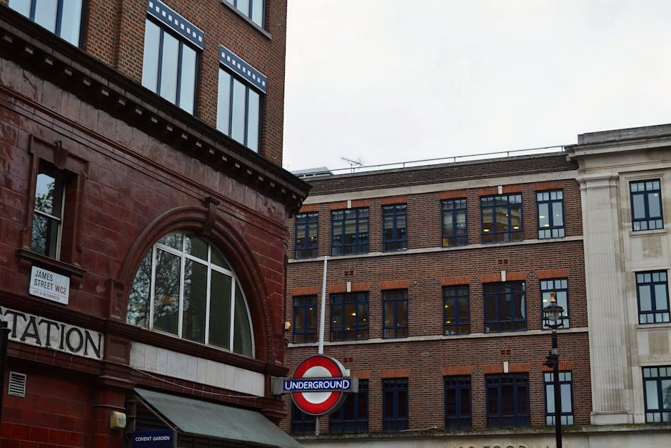 A view of the exterior of a multi-storey brick building in Covent Garden, showing windows with dark frames and a white stone section on the right side. In the foreground, part of a red-brick structure with a large arched window and decorative architectural elements is visible, along with a street sign that reads 'James Street WC2' and a Council sign indicating 'Underground' transport nearby. The sky is overcast, casting diffuse lighting over the scene, which is typical for a city environment involved in home relocation or moving activities, though no moving equipment or individuals are visible in this image.