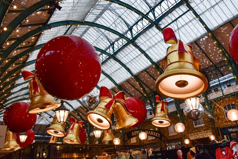 Inside a large indoor market with a high, curved glass ceiling, decorated with festive Christmas ornaments including large red baubles and golden bells tied with red ribbons. The bell-shaped light fixtures are suspended from the ceiling, illuminating the space, and there are vintage-style street lamps also hanging among the decorations. The market stall sign reads 'Apple Market,' with several small retail displays and clothing items visible below. The scene suggests preparations for holiday retail activity, reflecting the environment where Man With a Van Covent Garden might assist with home relocation or furniture transport within busy shopping venues during festive seasons, with a focus on efficient packing, loading, and moving logistics.