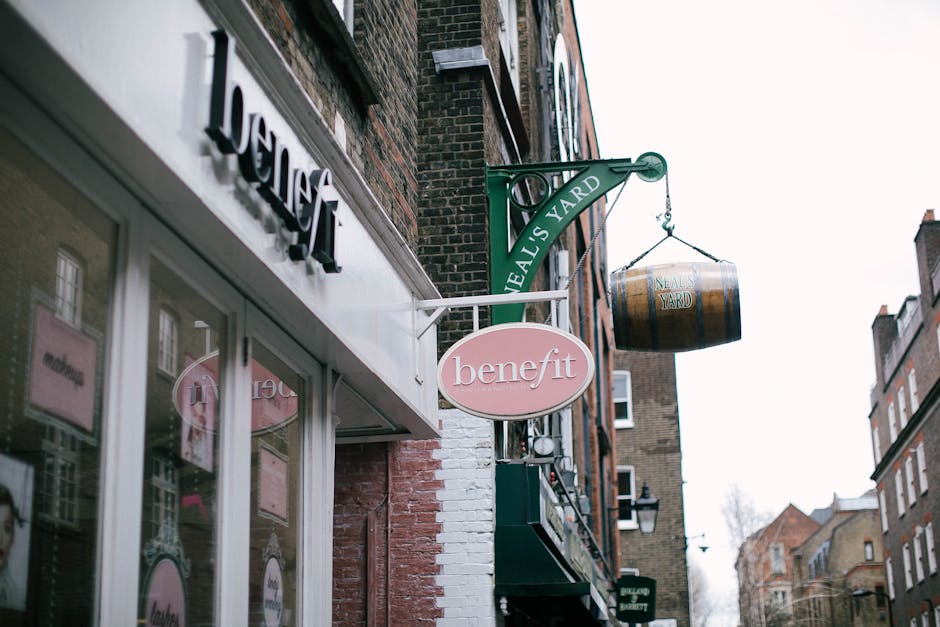 The image shows the exterior of a retail building situated on a narrow street in Covent Garden, featuring signage for a Benefit beauty store. Above the store, a green metal sign with the text 'Neals Yard' extends horizontally, supporting a wooden barrel-like object labeled 'Neals Yard,' hanging from a metal bracket. The building's facade includes a large glass display window reflecting nearby structures, with a partially visible pink Benefit sign mounted above the entrance. The street scene displays adjacent buildings constructed with brick, multi-story, with some windows showing interior light. The sky appears overcast, providing diffused natural light. This urban setting may be part of a home relocation or moving process involving the careful handling and transport of items through tight access points, consistent with services provided by Man With a Van Covent Garden.