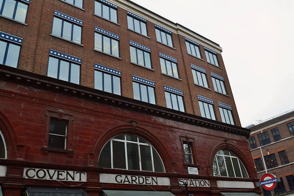 The image shows the exterior of a retail building situated on a narrow street in Covent Garden, featuring signage for a Benefit beauty store. Above the store, a green metal sign with the text 'Neals Yard' extends horizontally, supporting a wooden barrel-like object labeled 'Neals Yard,' hanging from a metal bracket. The building's facade includes a large glass display window reflecting nearby structures, with a partially visible pink Benefit sign mounted above the entrance. The street scene displays adjacent buildings constructed with brick, multi-story, with some windows showing interior light. The sky appears overcast, providing diffused natural light. This urban setting may be part of a home relocation or moving process involving the careful handling and transport of items through tight access points, consistent with services provided by Man With a Van Covent Garden.