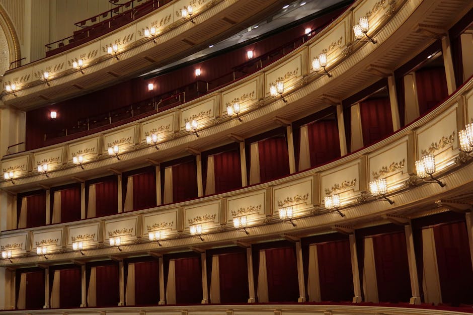 Interior view of the upper tiers inside the Royal Opera House in Covent Garden, showing rows of private boxes with cream-colored panels and gold decorative accents. Each box is separated by dark wooden partitions, and small, ornate wall-mounted lamps illuminate the space with warm lighting. The balconies are lined with elaborate moldings and supported by decorative brackets, creating a layered, curved arrangement. The rich red theater curtains are partially visible behind the boxes, contributing to the elegant, classical ambiance. This setting depicts the interior environment where house removals and furniture transport may involve careful access through entry points or loading areas adjacent to the theater. The image emphasizes the detailed architectural features and lighting typical of historic performance venues, relevant for professionals planning a home relocation involving delicate or valuable objects associated with the theater.