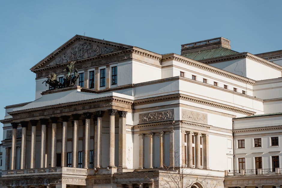 Photograph of the historic exterior of the Royal Opera House Covent Garden, showing its classical architectural features including tall Corinthian columns supporting a detailed frieze and triangular pediment adorned with sculptural reliefs. The building's white façade contrasts with a green-stained roof and a clear blue sky in the background. A bronze chariot sculpture with two horses is visible atop the pediment, emphasizing its grandeur. The image captures the front entrance area, which is used for house relocations and furniture transport during removals services. When performing packing and moving tasks, [COMPANY_NAME] often handles the careful loading and unloading of boxes and furniture onto external steps or into vans parked nearby, ensuring the preservation of this iconic structure during removal processes related to Covent Garden's cultural and historic environment.