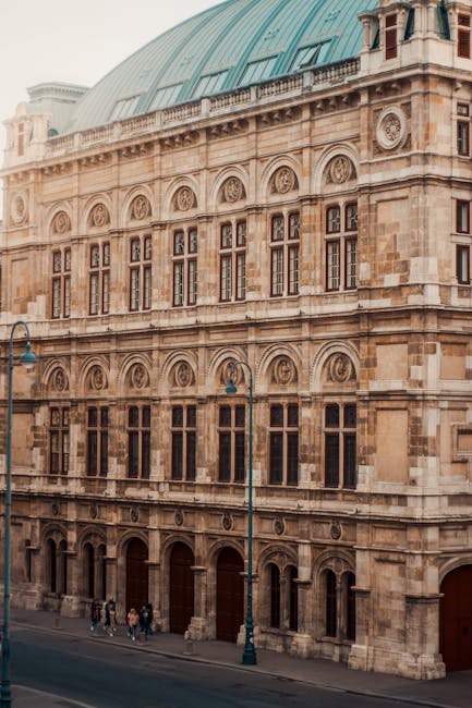 Interior view of the upper tiers inside the Royal Opera House in Covent Garden, showing rows of private boxes with cream-colored panels and gold decorative accents. Each box is separated by dark wooden partitions, and small, ornate wall-mounted lamps illuminate the space with warm lighting. The balconies are lined with elaborate moldings and supported by decorative brackets, creating a layered, curved arrangement. The rich red theater curtains are partially visible behind the boxes, contributing to the elegant, classical ambiance. This setting depicts the interior environment where house removals and furniture transport may involve careful access through entry points or loading areas adjacent to the theater. The image emphasizes the detailed architectural features and lighting typical of historic performance venues, relevant for professionals planning a home relocation involving delicate or valuable objects associated with the theater.
