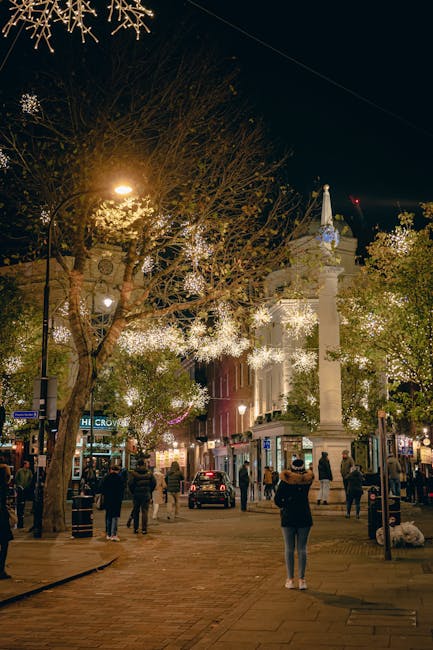 Inside a covered marketplace with a high glass ceiling, large decorative golden bells with red ribbons hang prominently at the entrance, creating a festive atmosphere. Below the bells, a curved black sign displays the words 'APPLE MARKET' in white lettering, attached to a metal archway. Surrounding the arch, there are multiple smaller lantern-style lights emitting a warm glow, along with red hanging ornaments and reflective silver globes, all contributing to the decorative display. In the background, shopfronts and additional lighting elements are visible along the marketplace interior. This scene captures a well-lit, festive space ideal for home relocation or furniture transport preparations, with a focus on decorative elements used to enhance the market's inviting ambiance. The photo provides a clear view of the marketplace's interior design and hanging decor, relevant to moving and packing logistics in busy commercial or retail environments, often associated with the area covered by Man With a Van Covent Garden and their removals services.