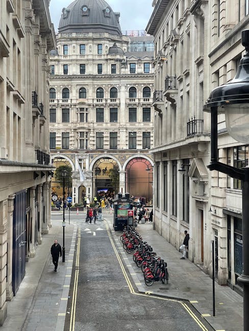 A narrow city street in Covent Garden with tall, ornate historic buildings on both sides, featuring detailed stone facades and balconies. The street is lined with parked red bicycles arranged neatly along the curb on the right side, while a black van is parked further into the scene, close to an arched passageway at the end of the street. Several pedestrians are walking along the pavement, some carrying bags, and others crossing the street. The sky is overcast, casting soft diffuse light on the scene. Visible in the distance are more buildings with classical architecture, and street lamps are mounted on the buildings. The scene appears to be in a busy urban area suitable for home relocation or furniture transport, with vehicles and pedestrians engaged in typical moving or loading activities. This setting aligns with the criteria for house removals and moving services, as provided by Man With a Van Covent Garden.