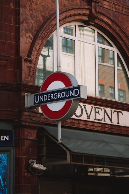 A close-up view of a London Underground sign mounted on a pole outside a building with red brick and arched windows, indicating Covent Garden station. The background shows part of a large window with white framing, reflecting nearby buildings, and a partial sign reading 'COVENT.' The scene is set during daylight, with natural light illuminating the area. This image exemplifies the urban environment where house removals and furniture transport activities may take place, especially in busy central London locations like Covent Garden, where careful logistics and adherence to permit rules are essential for safe and efficient moving processes as offered by Man With a Van Covent Garden.