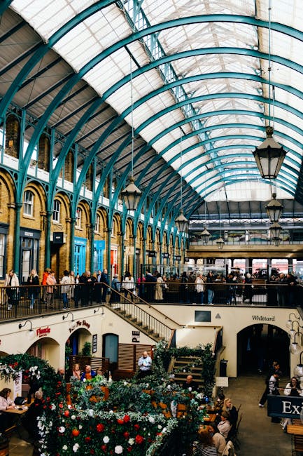 An indoor market hall with a large, curved glass and metal roof supported by teal-colored arches. The space is bustling with people walking along the wide central aisle and standing near the storefronts lining the perimeter, which have large windows and decorative brickwork. On the lower level, there are seating areas with tables and chairs, some occupied by shoppers or visitors. Potted plants and floral displays are arranged along the railings and near the seating areas, adding decorative greenery. The environment is well-lit with natural light filtering through the glass roof, complemented by hanging lantern-style lights. This setting, part of a busy marketplace or shopping venue, reflects a lively atmosphere suitable for socialising and browsing various shops, relevant to the context of home relocation and moving logistics such as loading or packing after a house move, as seen in the activities of people inside a well-organised, spacious indoor location like Covent Garden market, associated with professional removals service provider Man With a Van Covent Garden.