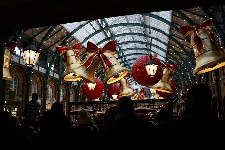 Inside a covered shopping arcade with a glass and metal arched ceiling decorated for the Christmas season, large gold and red bell-shaped ornaments with red bows, and red spherical baubles are hanging from the ceiling, illuminated by attached lantern-style lights. The area is filled with people, some seated and others walking, suggesting a busy atmosphere during a holiday event. In the background, glimpses of storefronts and additional festive decorations are visible. The lighting is warm yet subdued, emphasizing the holiday decorations. This scene depicts a typical shopping environment involved in home relocation or furniture transport, as part of packing and moving preparations. Man With a Van Covent Garden, a professional removals service, may use such decorated locations for moving or logistical planning, with the image capturing the festive ambiance often associated with city-based house removals during the holiday season.